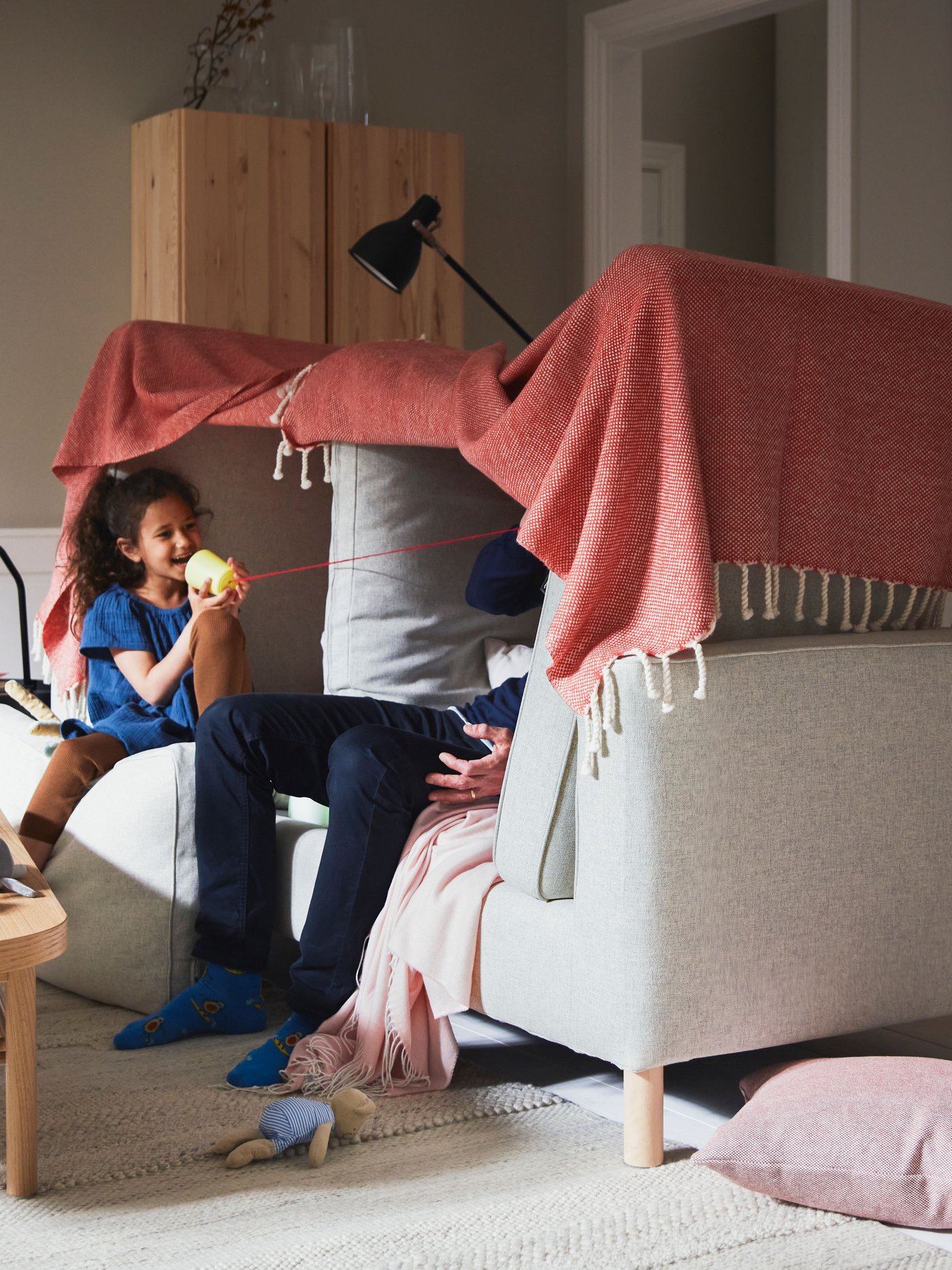 In a living room are a parent and a child playing in a beige sofa with pink pillows and a red blanket. Toys on the floor.