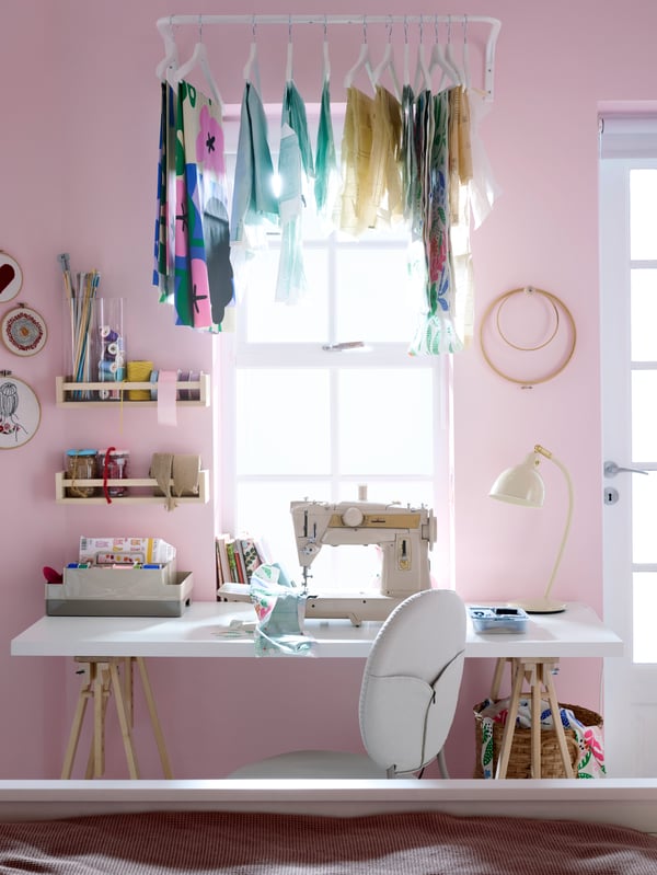 A desk with a sowing machine in front of a pink wall and window with bright light shining through. Textiles hanging above.