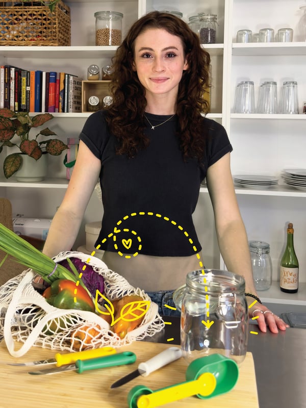A woman standing behind a kitchen counter. A net bag filled full of vegetables is placed on the counter in front of her.