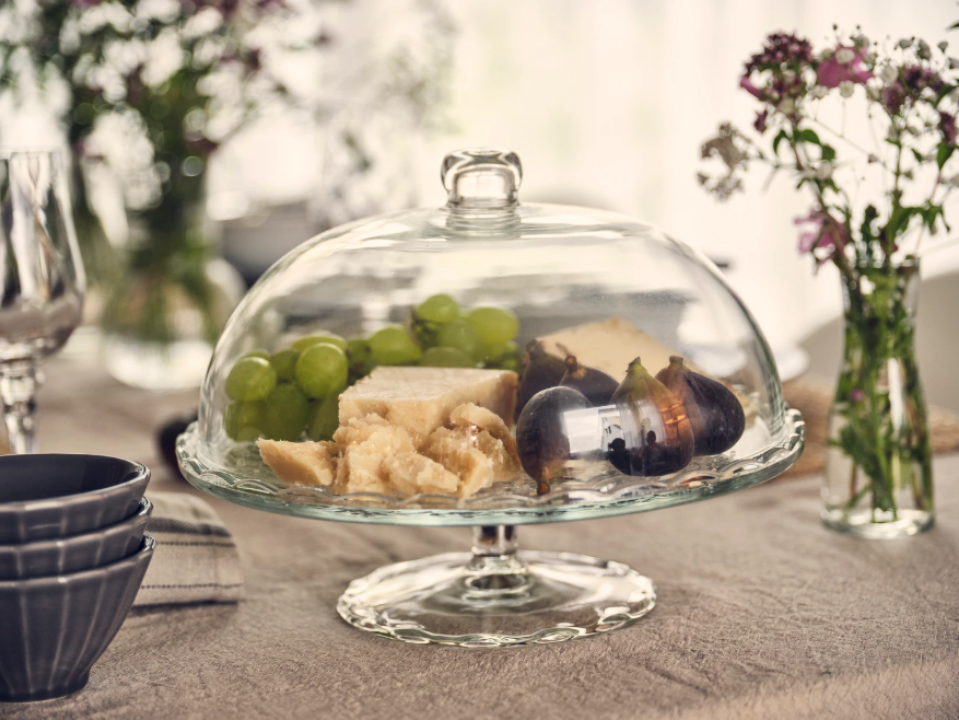 Käse, Trauben und Feigen unter einer Glashaube auf einer Glasplatte, im Hintergrund Geschirr und Blumen auf gedecktem Tisch.