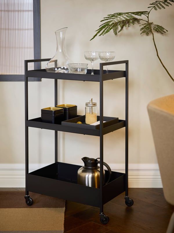 A hotel room has a black-coated steel, three-shelved NISSAFORS trolley with a carafe, glassware, tea tins and a vacuum flask.