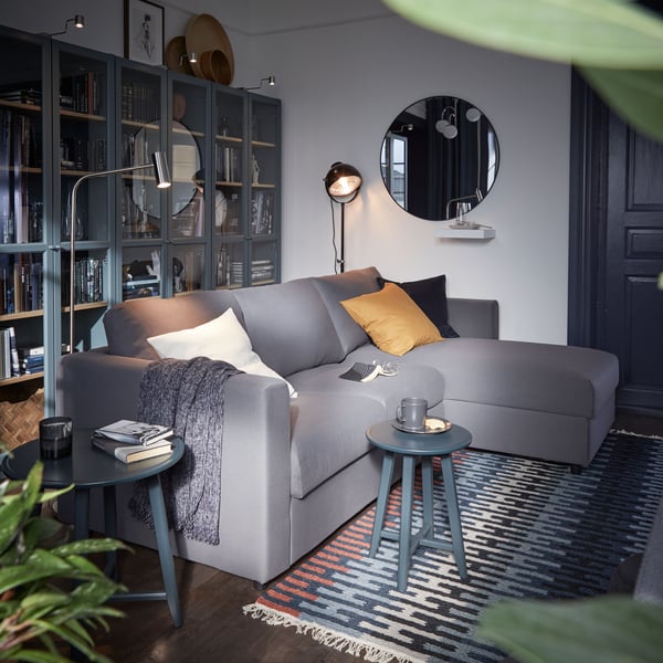 Modern eingerichtetes Wohnzimmer mit dunkelgrauem Ecksofa, gelbem Kissen, blauer Teppich, runder Spiegel und Regalwand mit Büchern