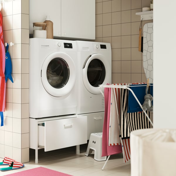 An ENHET pedestal, placed under a washing machine and drier, in a tiled laundry room with a drying rack.