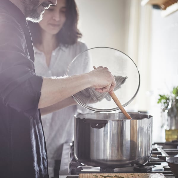 A man and a woman looking a the MIDDAGSMAT pot atop the stove with the man holding the pot lid and stirring the pot.