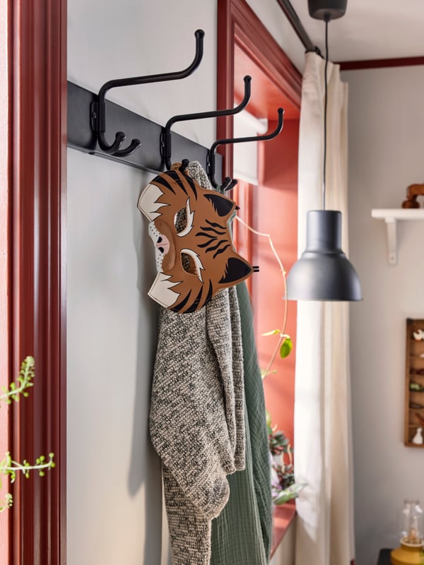 PINNIG rack with three hooks on white wall, with a mix of clothes and SKOGSDUVA mask hanging. Black lamp in window behind.