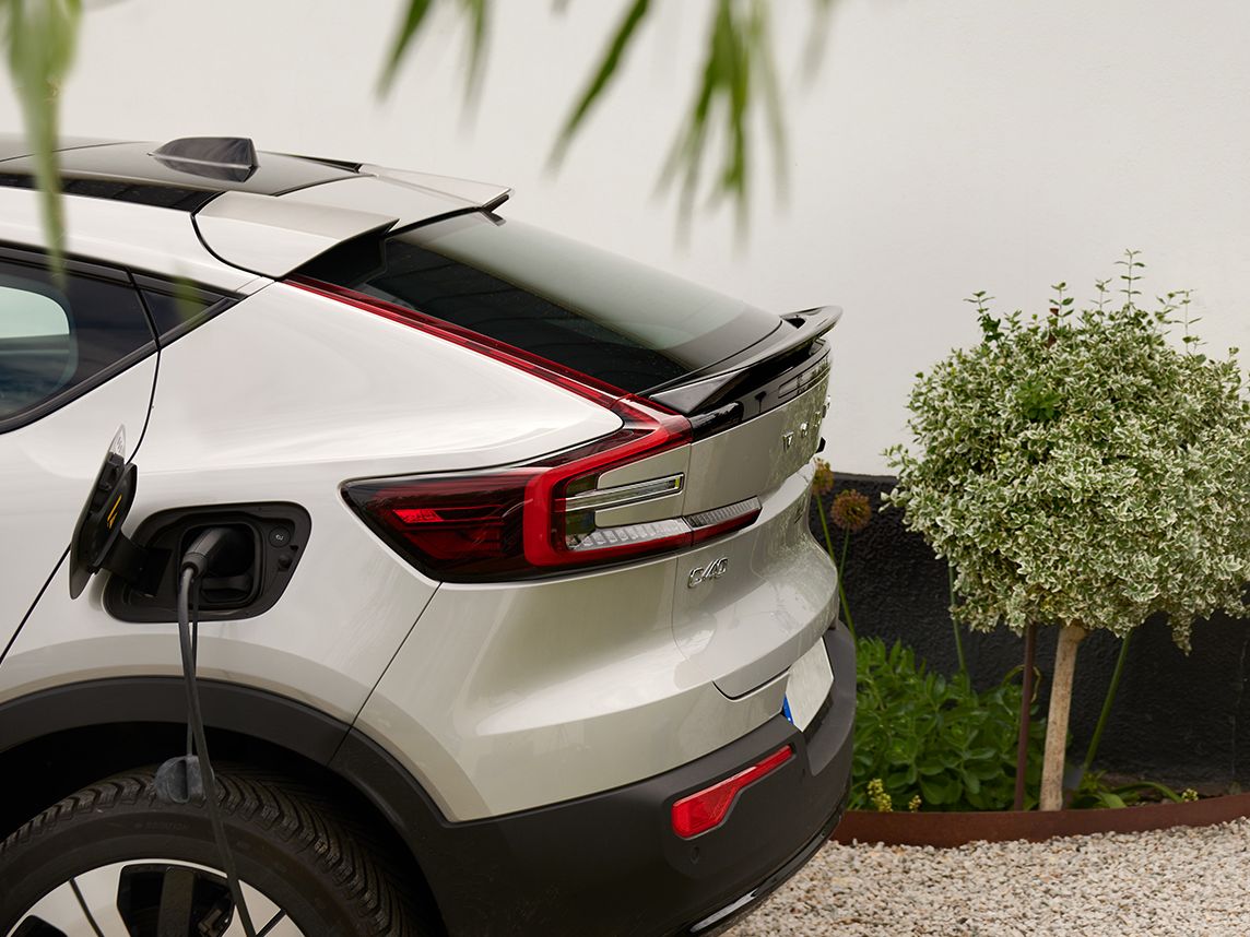 Silverfärgad elbil står parkerad vid sidan av ett vitt hus med svart tak och mörkgrå takpannor. Bilen är ansluten till en väggmonterad laddstation. Framför huset finns grusgång, gröna växter i krukor och en liten trädgård med buskar.