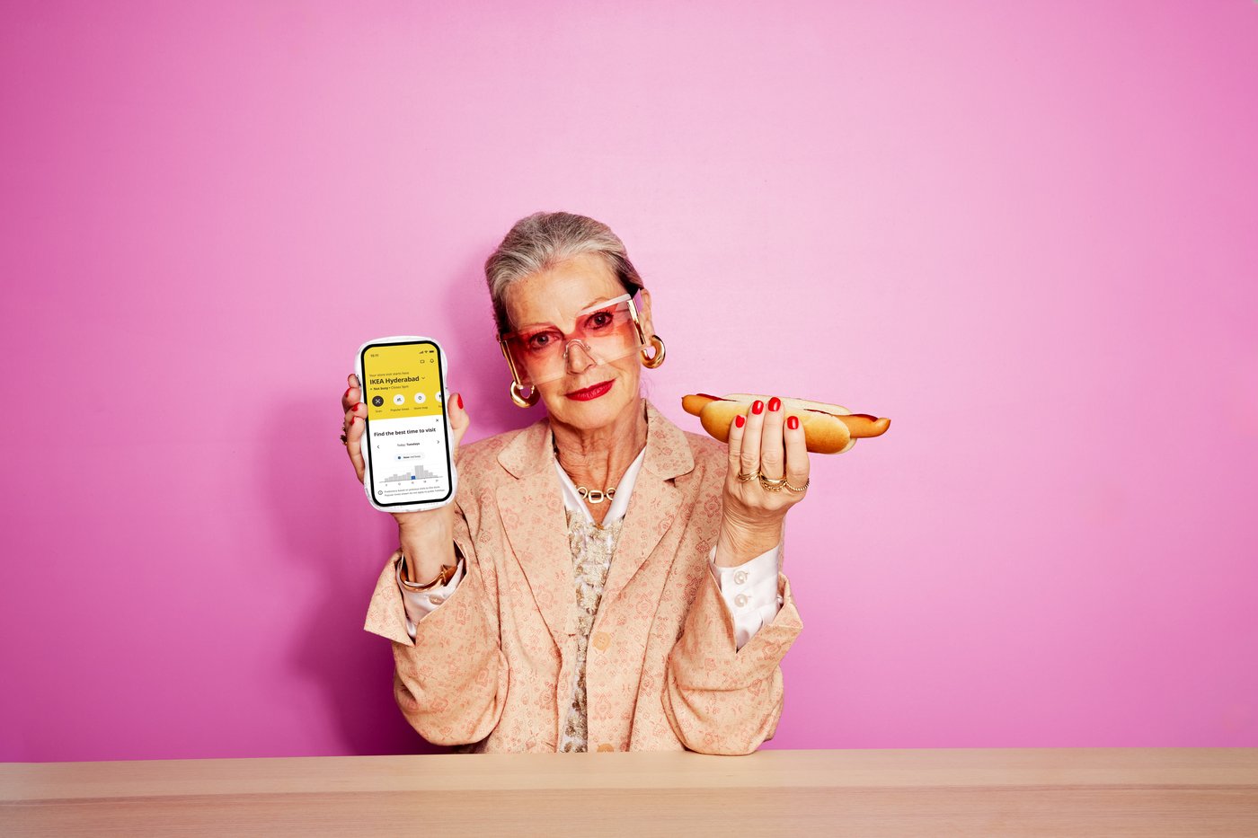 Person hält ein Smartphone mit geöffneter IKEA App in der Hand und sitzt vor einem rosa Hintergrund an einem Tisch.