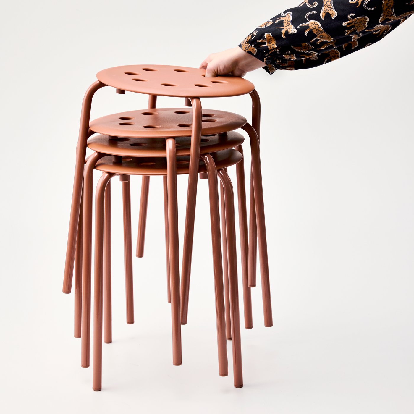 Una persona col·loca un tamboret MARIUS de color marró rogenc en una pila amb altres tamborets del mateix model. Se n'aprecia el disseny lleuger i la versatilitat.