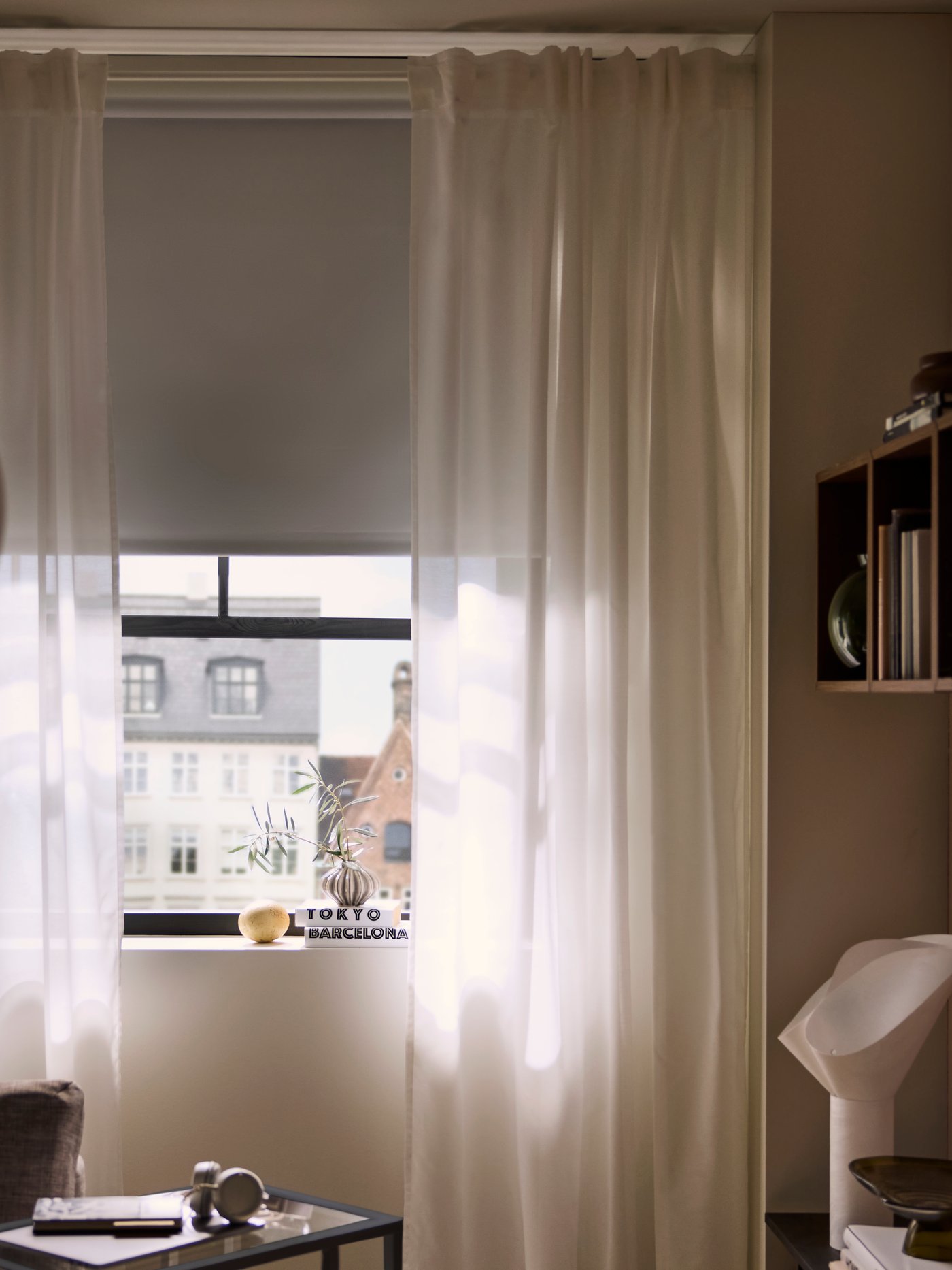 A sunlit window with a FRIDANS block-out roller blind and curtains partly shut, dimming the interior of the room inside.
