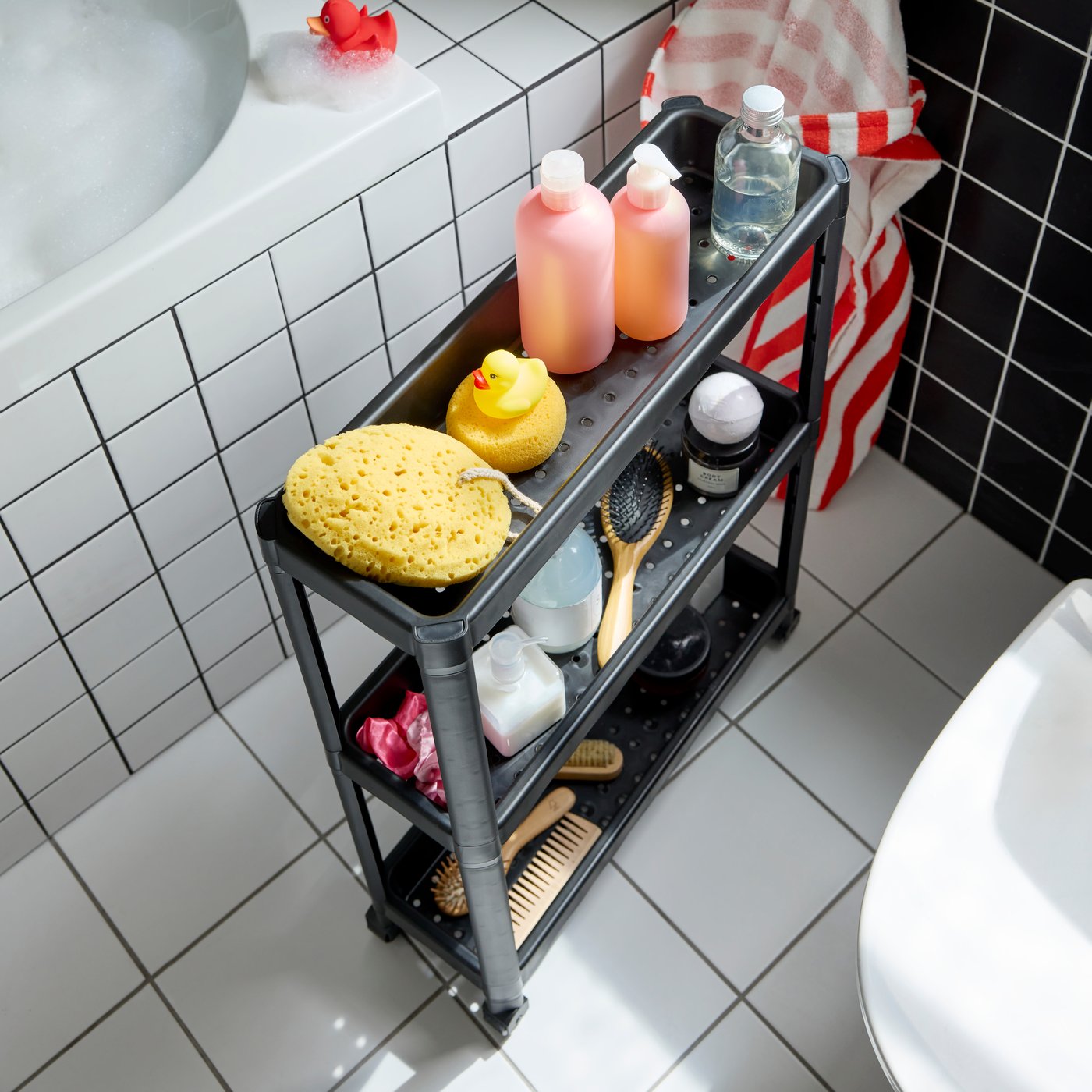 A black VESKEN trolley with three shelves filled with hair brushes, shampoo bottles and kid’s bath toys. White floor.