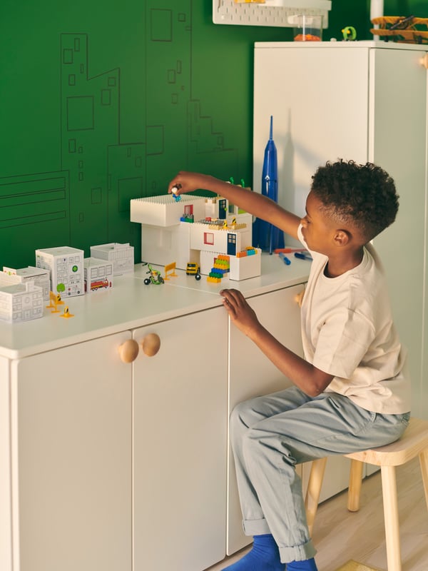 A boy sits on a stool, playing with a BYGGLEK LEGO brick set placed on a white SMÅSTAD cabinet-and-wardrobe combination.