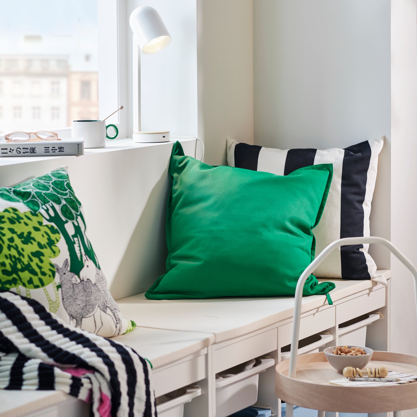 A cushion in a green GURLI cover on top of two white TROFAST units beside a window sill holding a white ENSMÄRKE desk lamp.