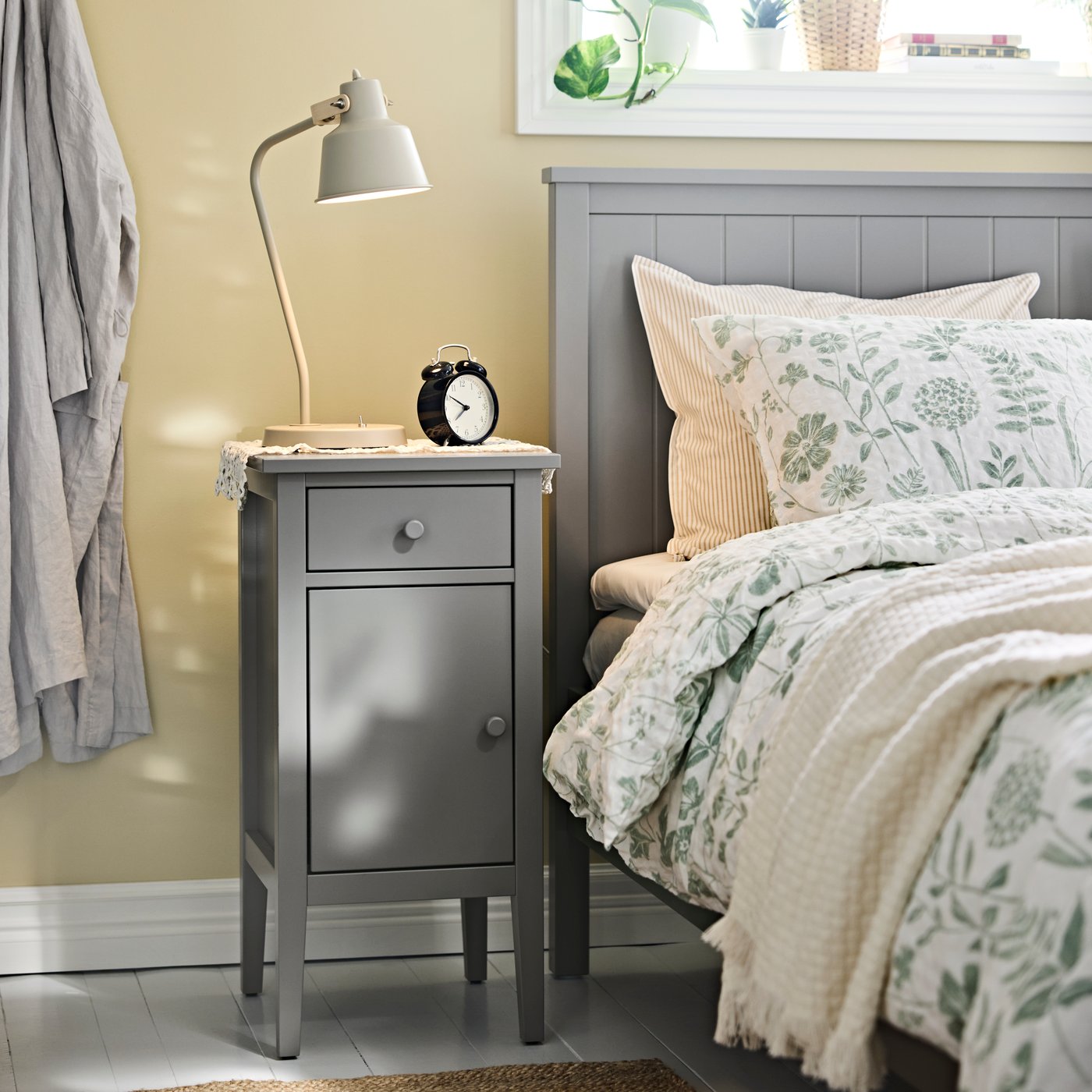 A bedroom with a GULLABERG bedside table next to a dressed bed, with a lamp, clock and a robe hanging next to it.