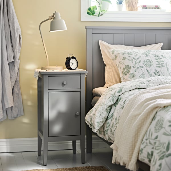 A bedroom with a GULLABERG bedside table next to a dressed bed, with a lamp, clock and a robe hanging next to it.