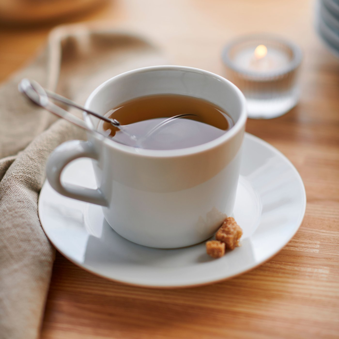 A tea infuser is placed in a white IKEA 365+ cup on a bamboo surface. A small crumble of raw sugar is placed on the saucer.
