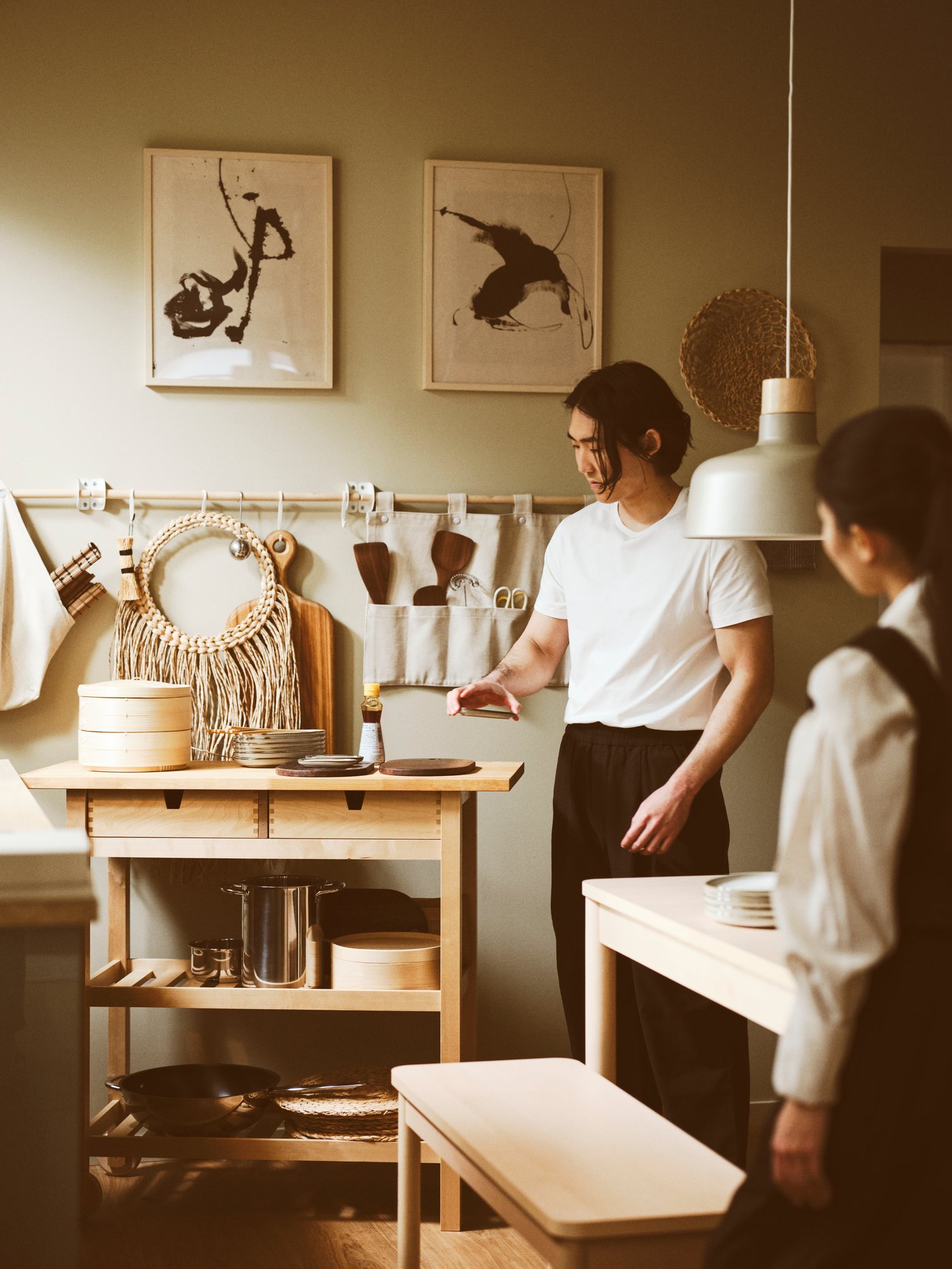 A kitchen featuring two people, a FÖRHÖJA kitchen trolley, BUNKEFLO pendant lamp, and a RÖNNINGE extendable table.