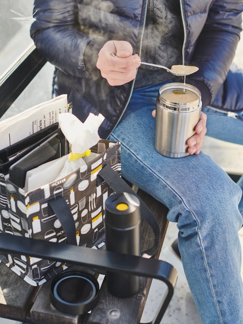 A man sitting by a FLADDRIG lunch bag on a public bench, eating with a spoon out of an EFTERFRÅGAD food vacuum flask.