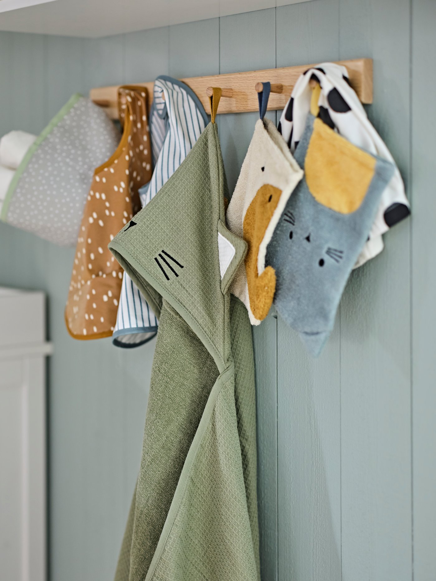 A wooden rail with hooks holds a number of different items from GULDVÄVARE collection including wash mits, bibs and a towel.