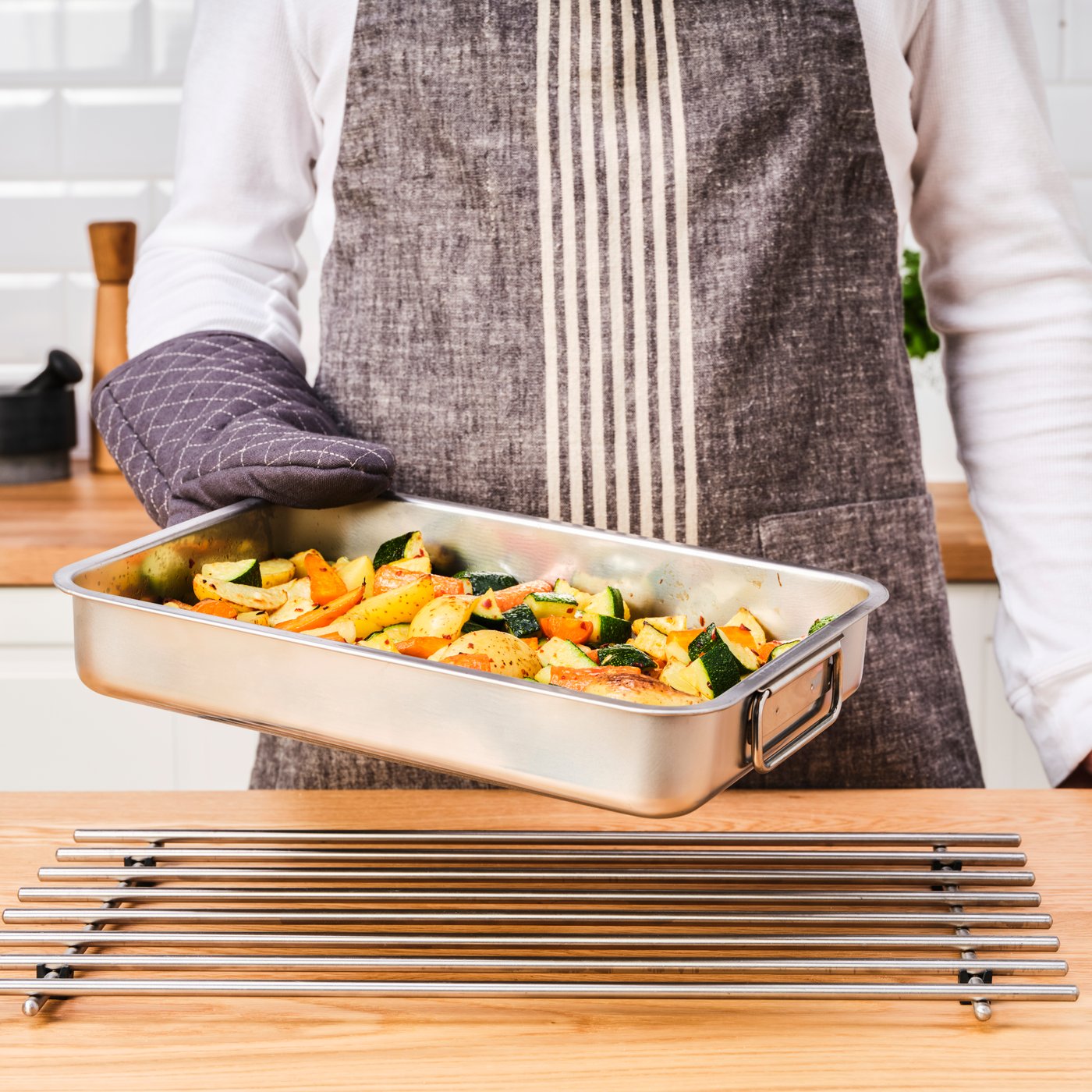 Eine Person mit einer Schürze hält mit einem Ofenhandschuh eine KONCIS Ofenform aus Edelstahl, in der sich Gemüse befindet, in der Hand. 