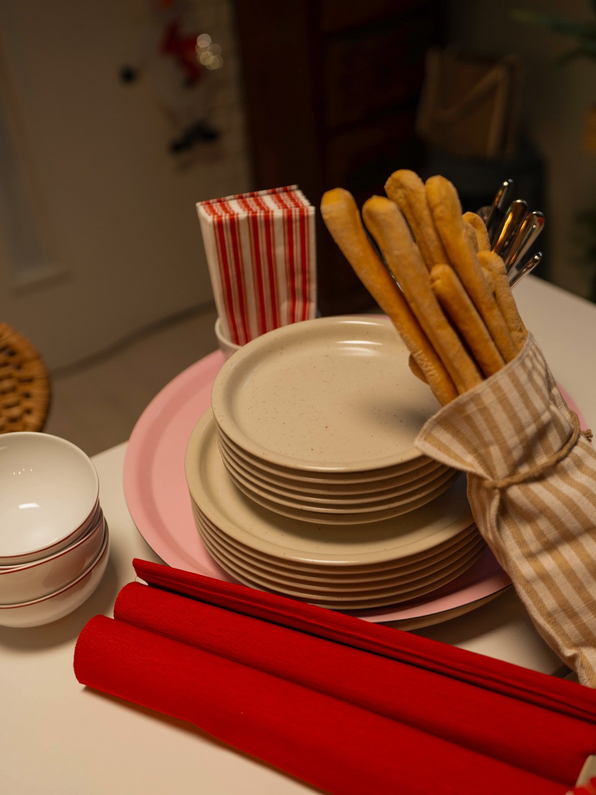 Una pila di piatti beige DAGGASTER, grissini avvolti in un tovagliolo di stoffa a righe e tovaglioli di carta rossi, pronti per apparecchiare la tavola delle feste.