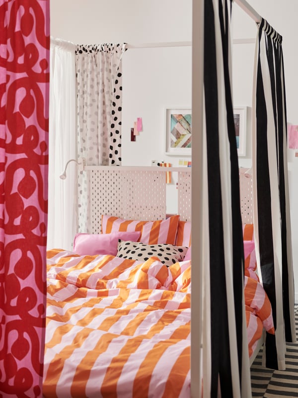 Una cama con dosel VITARNA blanca vestida con ropa de cama SLÅNHÖSTMAL y con la tela precortada VARGTÖREL y FLUGBLOMSTER alrededor.