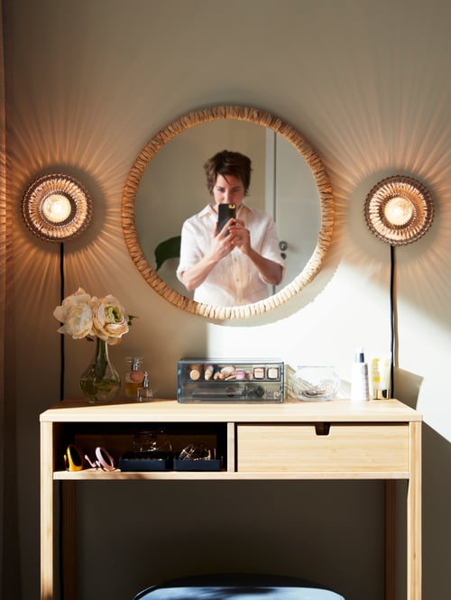 Bedroom featuring a person standing in front of a rectangular dressing table made of bamboo, placed under a round mirror.