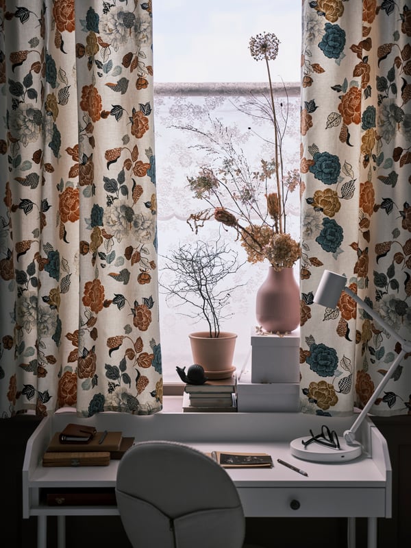 Curtains made with flower patterned TROLLMAL fabric in a window above a white HAUGA desk with a beige swivel chair.