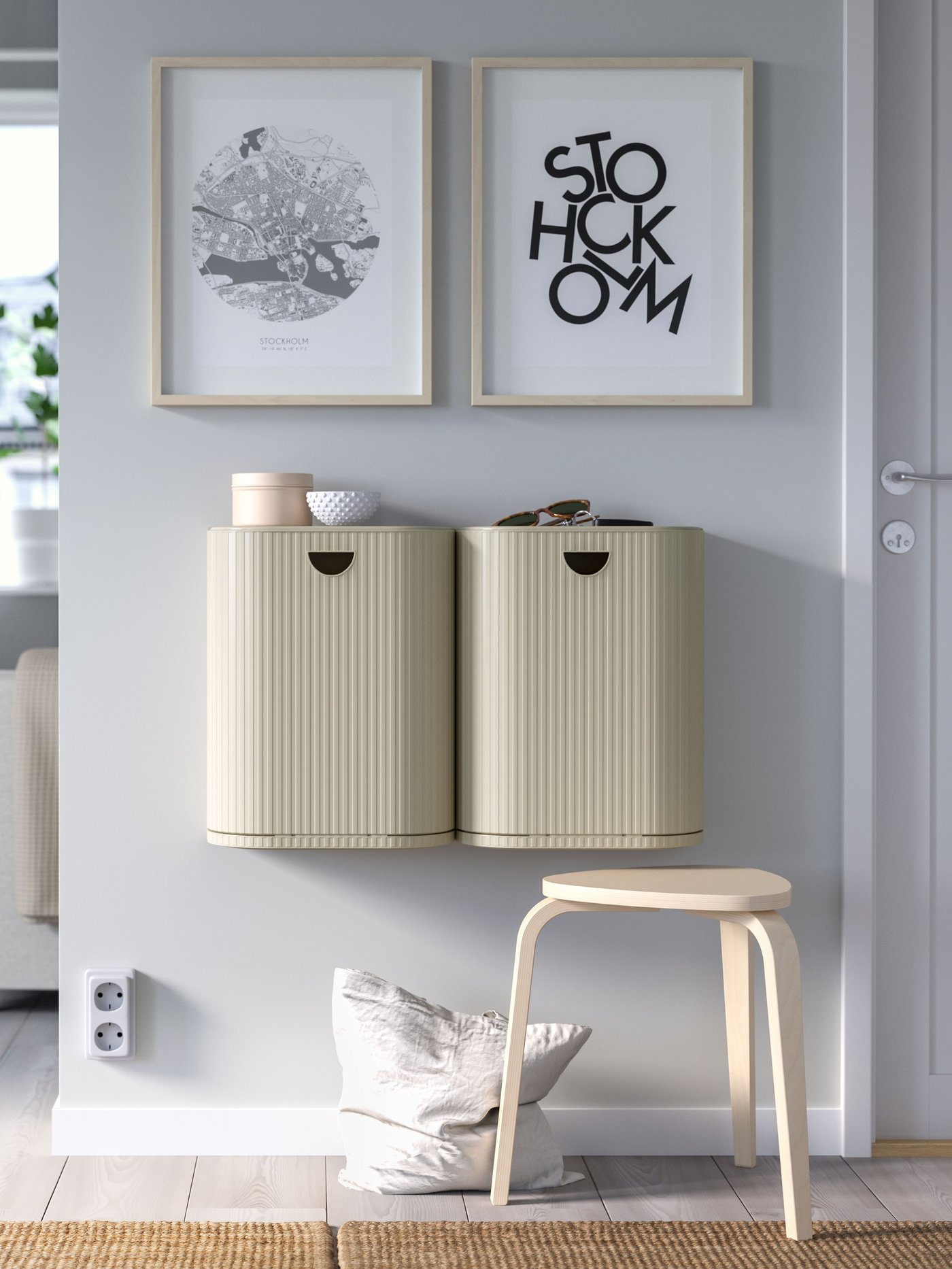 Två väggmonterade beige GÅRDSBUTIK med ribbad yta, dekor ovanpå, en pall, kudde på golvet och två tavlor – en abstrakt och en med texten "STOCKHOLM".