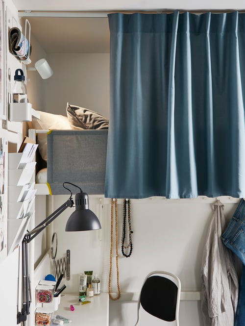 Es ist ein Schlafzimmer mit einem VITVAL Hochbettgestell mit Arbeitsplatte in Weiß/Hellgrau zu sehen. Die Arbeitsplatte wird hier als Frisiertisch genutzt. Vor der Liegefläche hängt ein Vorhang, um für Privatsphäre zu sorgen. 