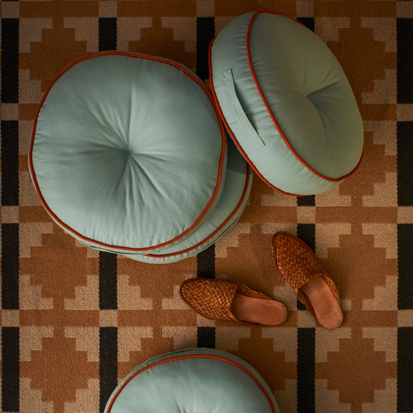 Empat kusyen lantai GOKVÄLLÅ pada permaidani di sebelah sepasang selipar.