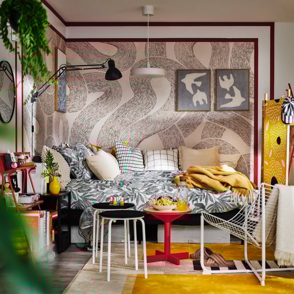 Two MARIUS stools and a red side table by a SLATTUM upholstered bed with BROKDRACENA patterned bed linen and cushions on top.