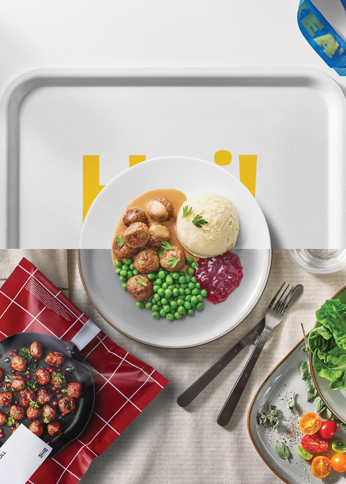 그레이 트레이 위에 놓인 접시에 스웨디시 미트볼과 매시드 포테이토, 그린피스, 링곤베리 잼이 담겨 있다. 옆에는 포크와 나이프가 놓여 있으며, 한쪽에는 미트볼을 굽는 팬과 샐러드 접시 이미지