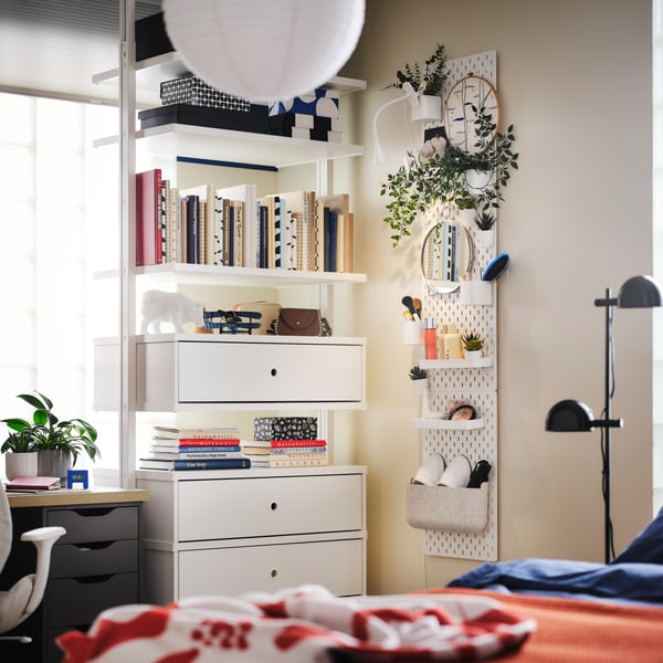 A white ELVARLI drawer unit, shelving and posts in a bright bedroom holding books and decorative items and by a pegboard.