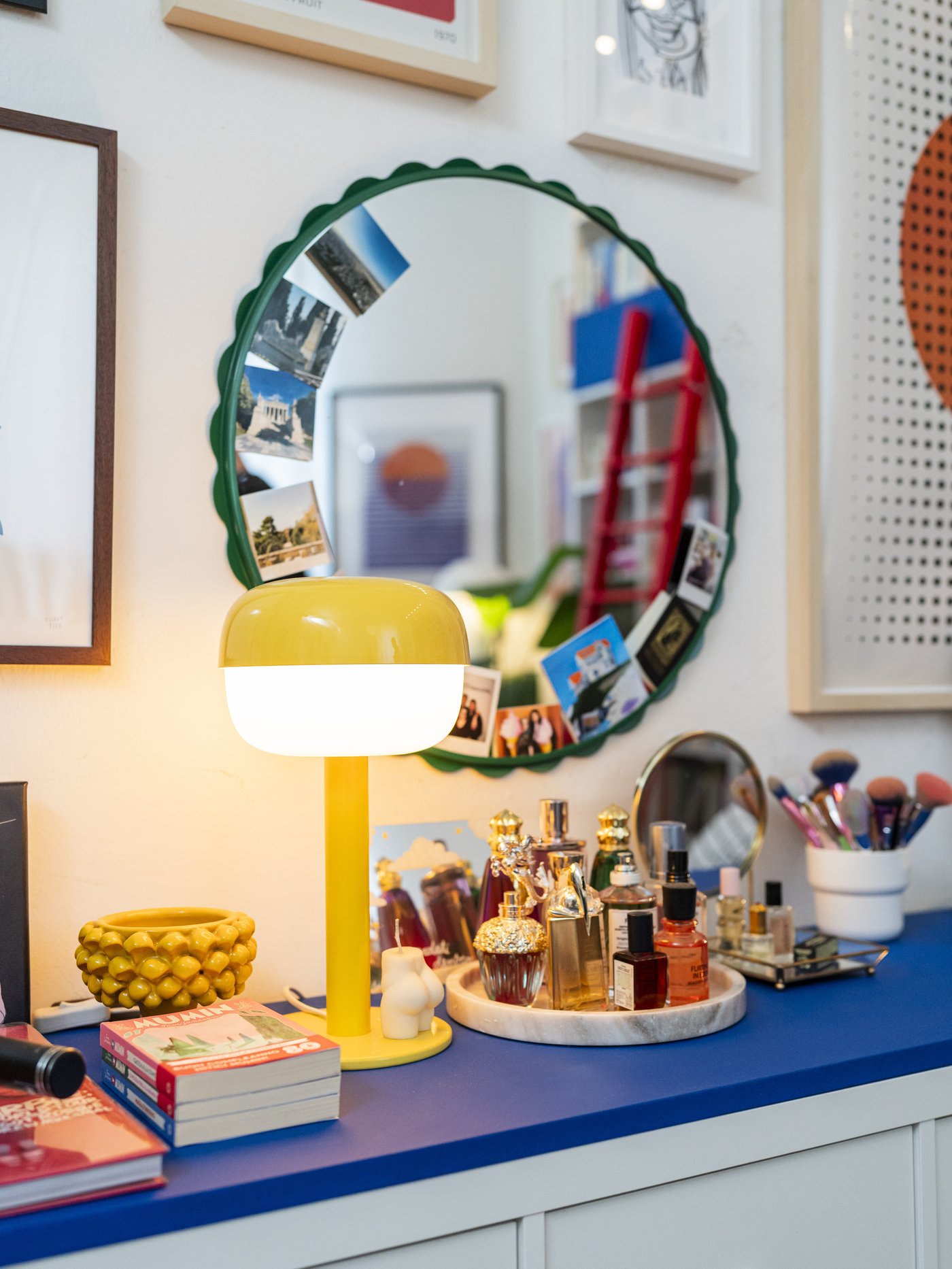 Lampada da tavolo gialla BLÅSVERK accesa su una superficie blu, circondata da libri, profumi e accessori davanti a uno specchio rotondo decorato con foto.
