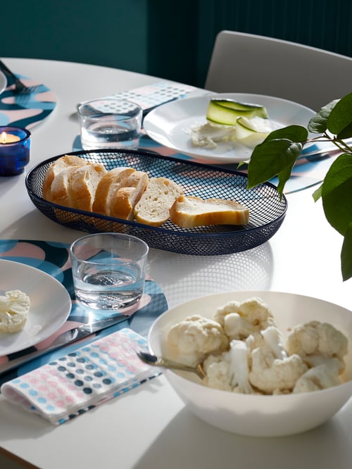 Table with colourful place mats, blue bread basket, white dinnerware, and white tempered glass bowl with cauliflower.