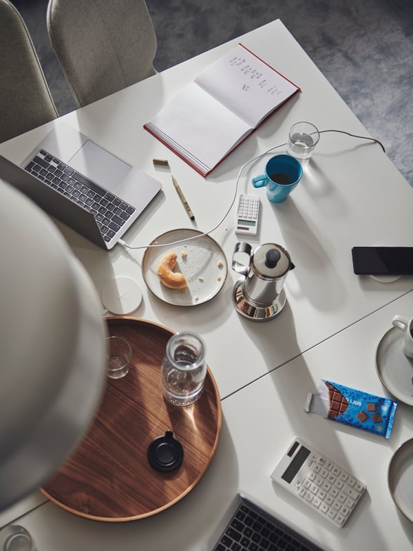 Mesa formada por duas secretárias TROTTEN em branco, lado a lado, com acessórios de escritório e para a pausa para café espalhados.