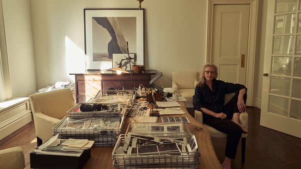 Annie Leibovitz sentada numa cadeira junto a uma mesa com muitas coisas na sua casa.