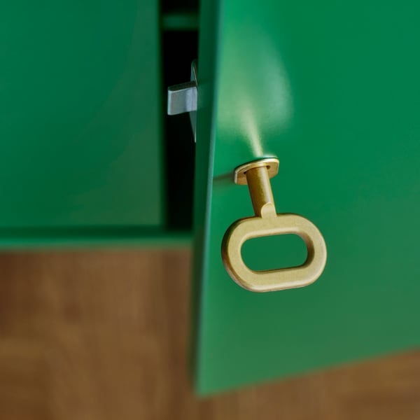 A close up of an oval-shaped brass key in the keyhole of a green STOCKHOLM 2025 cabinet with two lockable doors.
