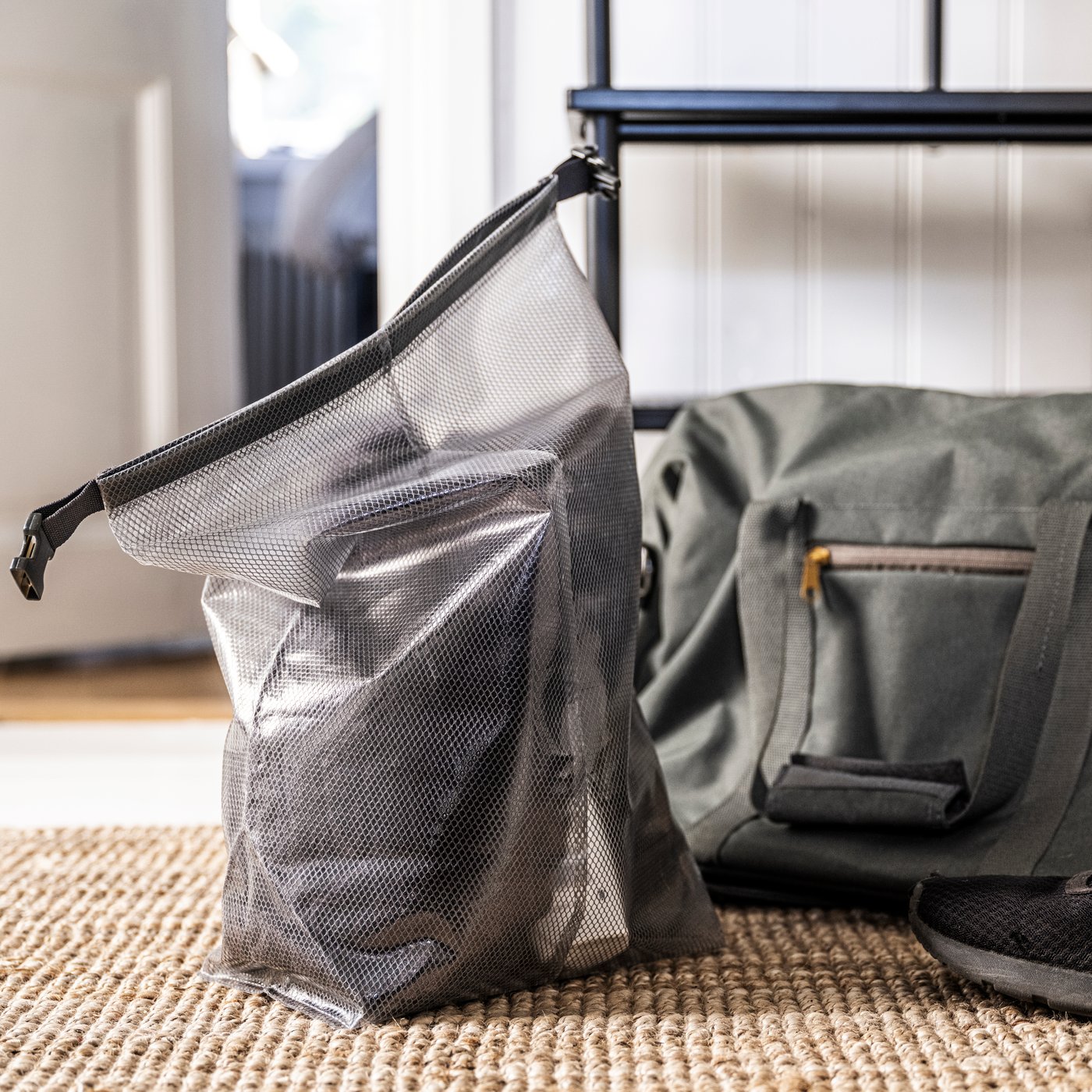 Průhledný uzavíratelný sáček IKEA RENSARE s uloženými botami stojí na jutovém koberci vedle zelené sportovní tašky.