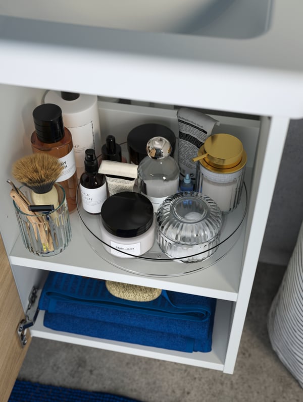 A white bathroom vanity with open doors has a KLIPPKAKTUS storage turntable on the top shelf, with bathroom products on it.