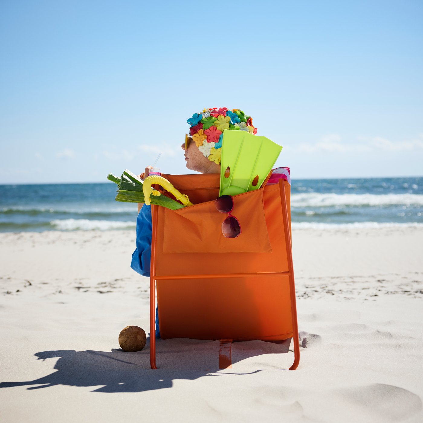 A beach setting features a multicolour STRANDÖN sun lounger with a person enjoying a drink and wearing a floral hat.