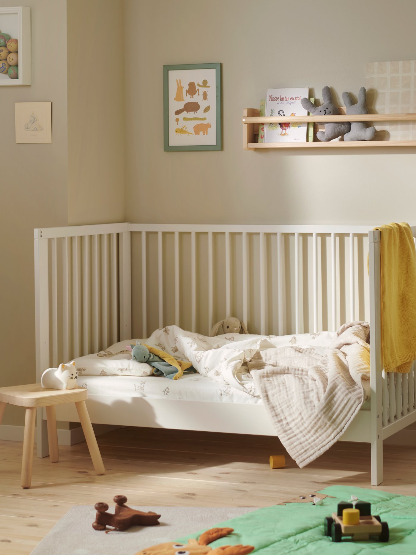 A GULLIVER cot in white wood in a neutral nursery, with bedding and a yellow towel, and toys on a green play mat below.