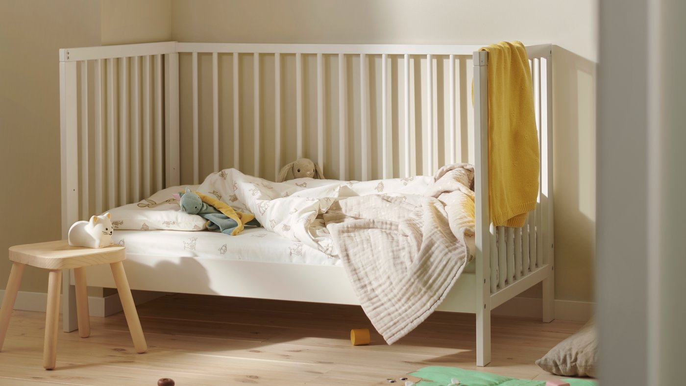 Una culla GULLIVER in legno bianco in una cameretta neutra, con biancheria da letto, un asciugamano giallo e alcuni giocattoli su un tappetino da gioco verde in basso.