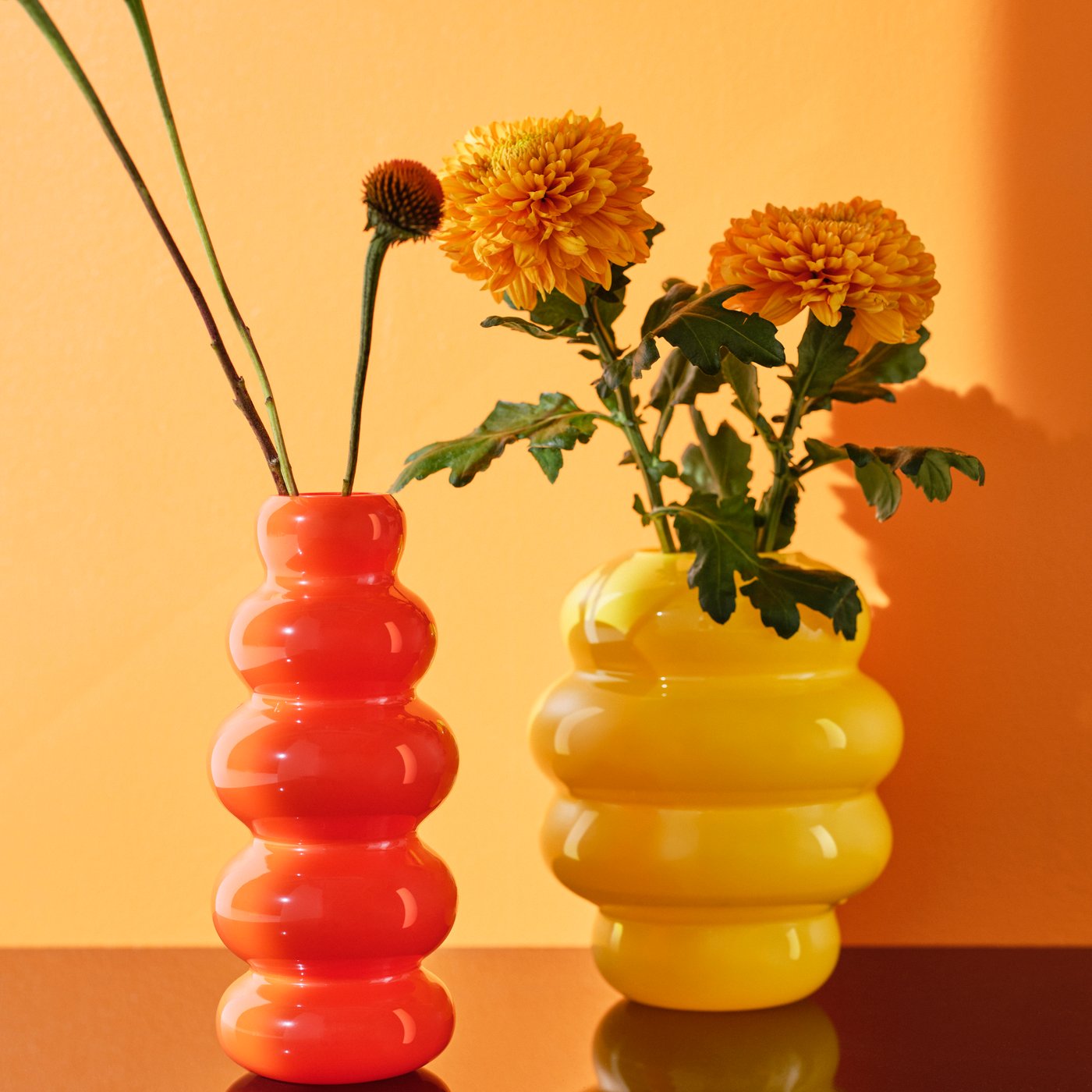 One orange and one yellow KÄLLARHALS vase with flowers in them are placed on a glossy surface that reflects the vases.