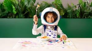 Child in astronaut costume eating at a creatively decorated table with drawings and markers.
