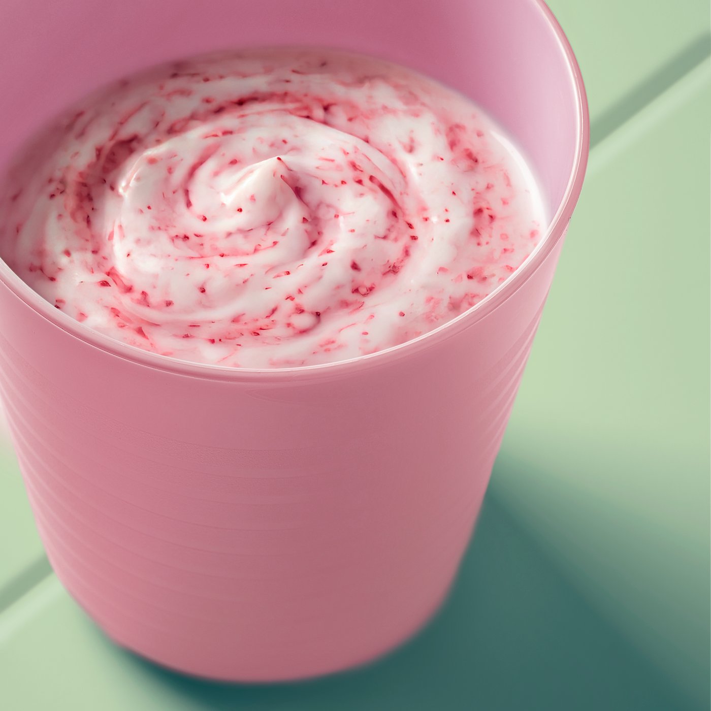 Close-up of a pink kid’s KALAS mug filled with creamy pink smoothe, shown on a light green table outdoors.