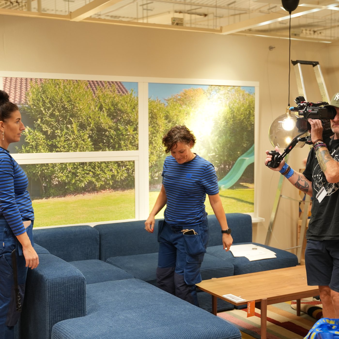 En optagelse fra "Hvem vinder hjem?" TV-programmet, hvor to indrettere rykker rundt på en sofa i et IKEA varehus, mens en kameramand filmer processen.