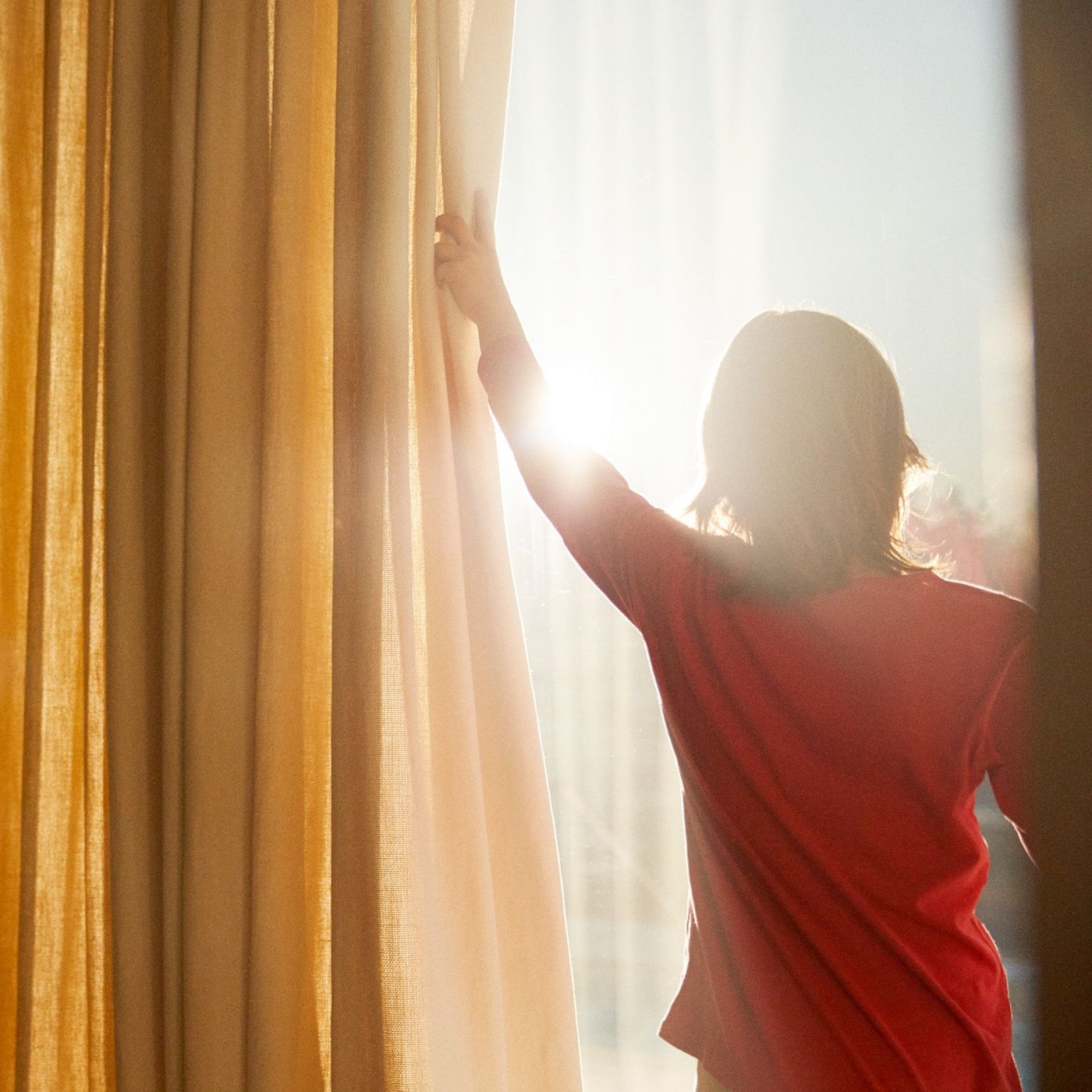 En person i röd tröja drar undan en gardin framför ett fönster och släpper in solljus i rummet.