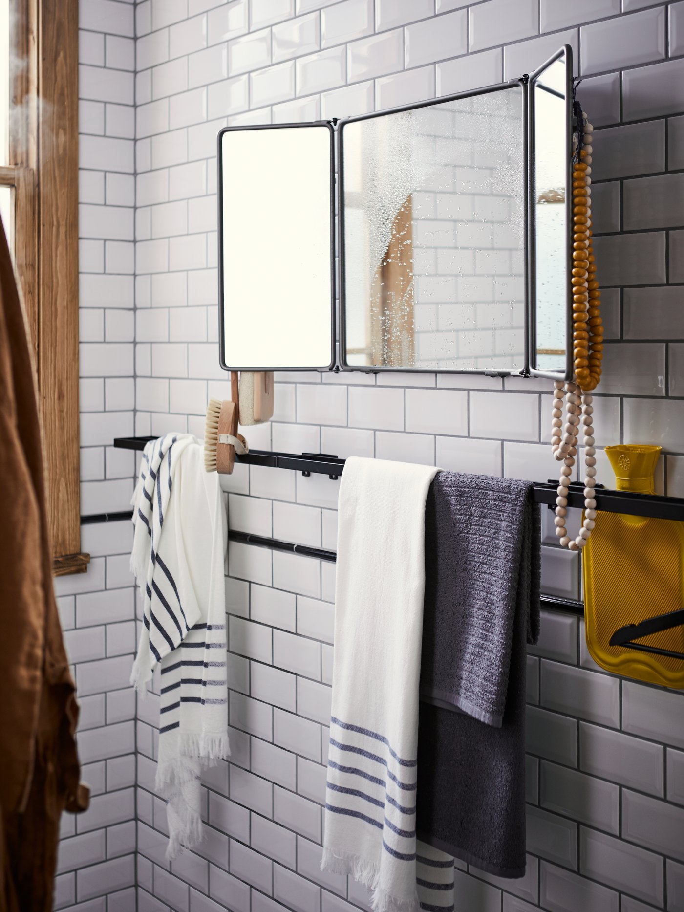 SKOGSVIKEN black towel rail with towels against a white tile bathroom wall with a SYNNERBY tri-fold mirror above.