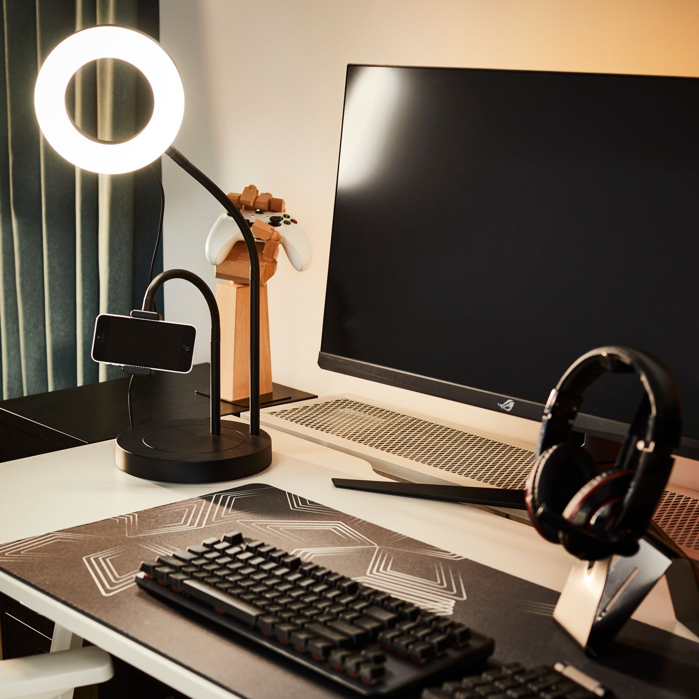 A LÅNESPELARE ring light with phone holder on a desk beside a monitor and keyboard and a pair of headphones on a stand.